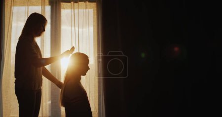 Téléchargez les photos : Silhouette d'une mère peignant les cheveux de ses filles à la fenêtre. - en image libre de droit