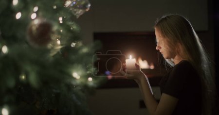 Téléchargez les photos : Une fille avec une bougie à la main est assise près du sapin de Noël. En arrière-plan, un feu brûle dans une cheminée. La veille de Noël. Vue latérale - en image libre de droit