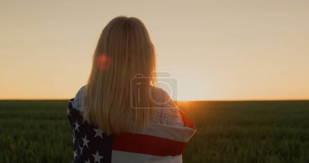 Foto de Una mujer con una bandera americana sobre sus hombros ve el sol ponerse sobre un campo de trigo - Imagen libre de derechos
