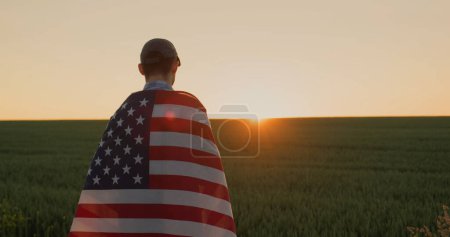 Foto de Joven agricultor masculino con bandera de EE.UU. en sus hombros mira al amanecer sobre el campo de trigo. - Imagen libre de derechos