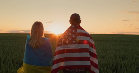 Foto de Un hombre y una mujer con las banderas de Ucrania y los Estados Unidos se paran uno al lado del otro y miran el amanecer sobre un campo de trigo.. - Imagen libre de derechos