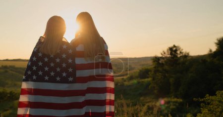 Téléchargez les photos : Une mère avec une adolescente avec un drapeau américain sur les épaules regarde le coucher du soleil sur une vallée pittoresque. - en image libre de droit