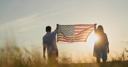 Foto de Un hombre y una mujer levantan la bandera de Estados Unidos sobre un campo de trigo al atardecer. concepto del día de independencia de EE.UU. - Imagen libre de derechos