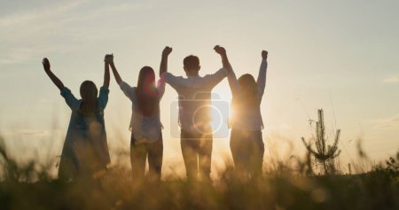 Foto de La silueta de un grupo de amigos - levanten las manos contra el telón de fondo del sol poniente. Trabajo en equipo y concepto de éxito. - Imagen libre de derechos