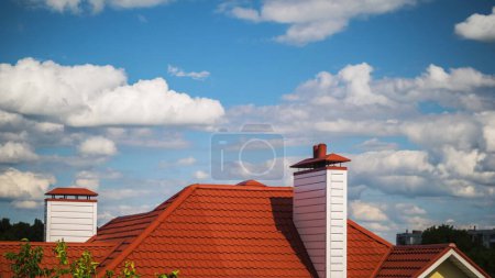 Foto de El techo de la casa está hecho de azulejos rojos, por encima de la casa hay un cielo azul con nubes en un día claro de verano. - Imagen libre de derechos