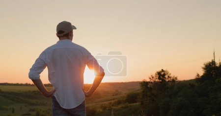 Téléchargez les photos : Un jeune agriculteur confiant se tient devant une campagne pittoresque au coucher du soleil. Vue de l'arrière. - en image libre de droit