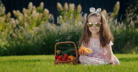 Téléchargez les photos : Une petite fille en lunettes de soleil est assise sur une pelouse verte près d'un panier de légumes - en image libre de droit