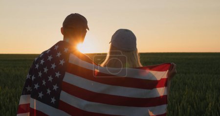 Photo for A man and a woman with the US flag on their shoulders look at the sunrise over a field of wheat. - Royalty Free Image