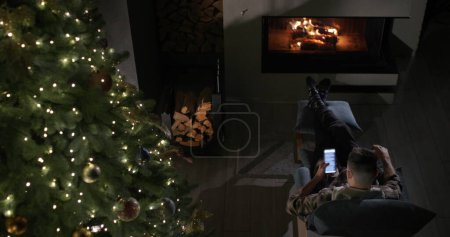 Téléchargez les photos : Un jeune homme utilise un smartphone. Il est assis dans un fauteuil près de la cheminée et un grand sapin de Noël. Vue d'en haut. - en image libre de droit