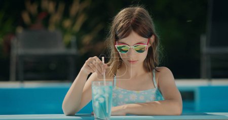 Foto de Retrato de una linda niña en gafas de sol al lado de la piscina. Muy cerca hay un cóctel fresco. - Imagen libre de derechos