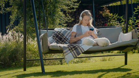 Téléchargez les photos : Une adolescente se repose sur une balançoire de jardin, à l'aide d'un smartphone - en image libre de droit
