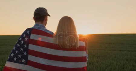 Foto de Pareja joven con bandera americana sobre sus hombros mirando hacia adelante en el campo de trigo al atardecer. - Imagen libre de derechos