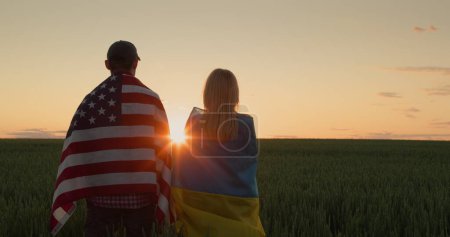 Foto de Pareja feliz con las banderas de Ucrania y los EE.UU. se paran uno al lado del otro y miran el amanecer sobre un campo de trigo. - Imagen libre de derechos