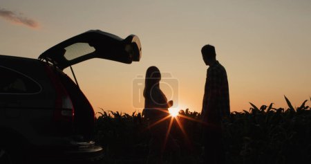 Foto de Una joven pareja admira la puesta de sol sobre el campo, de pie cerca de su coche. - Imagen libre de derechos