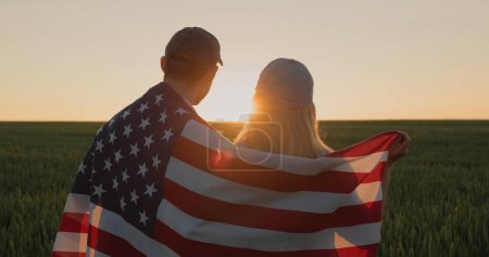 Foto de Pareja joven con bandera americana sobre sus hombros mirando hacia adelante en el campo de trigo al atardecer. - Imagen libre de derechos