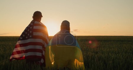 Foto de Pareja con las banderas de Ucrania y los EE.UU. de pie al lado del otro y mira la puesta de sol sobre un campo de trigo. - Imagen libre de derechos