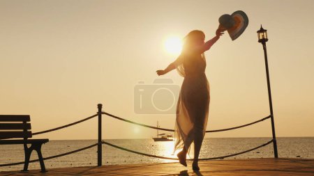 Photo for A woman meets the dawn at sea. Emotionally spinning with a hat in his hand on the pier. Dream of travel concept. - Royalty Free Image