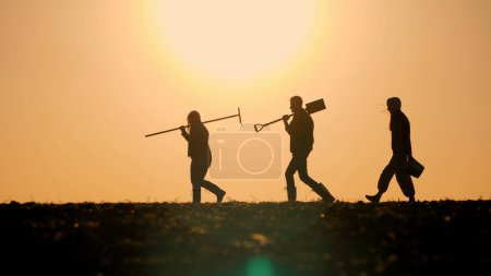 Foto de Una familia de agricultores con equipo de trabajo camina a través de un campo contra el telón de fondo de una hermosa puesta de sol. - Imagen libre de derechos