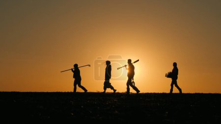 Téléchargez les photos : Une famille d'agriculteurs avec de l'équipement marche à travers le champ. Silhouettes au coucher du soleil. - en image libre de droit