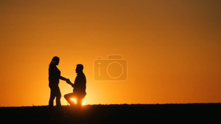 Téléchargez les photos : Silhouettes d'un homme et d'une femme au coucher du soleil. Un homme est à genoux devant une femme. Proposition de mariage. - en image libre de droit
