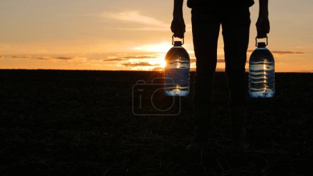 Foto de Un hombre sostiene dos botellas de agua potable, se para en un campo al atardecer. - Imagen libre de derechos