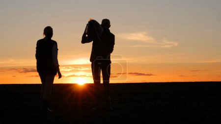 Two farmers are standing in a field at sunset. A man holds a bag on his shoulder, next to his wife.