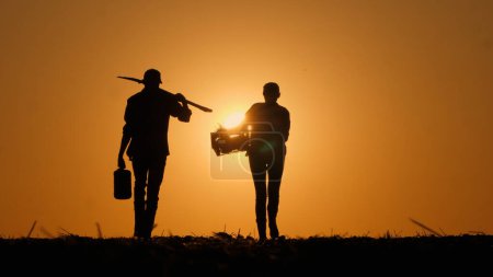 Silhouettes of two farmers walking across a field with working equipment towards the sunset. High quality photo