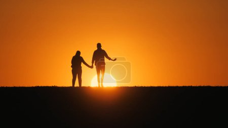 Téléchargez les photos : Tenant la main, un jeune couple marche ensemble à travers un champ, se déplaçant vers le soleil couchant - en image libre de droit