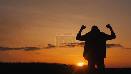 Téléchargez les photos : Silhouette d'un jeune homme dans un geste de victoire. Debout dans un champ au coucher du soleil. - en image libre de droit