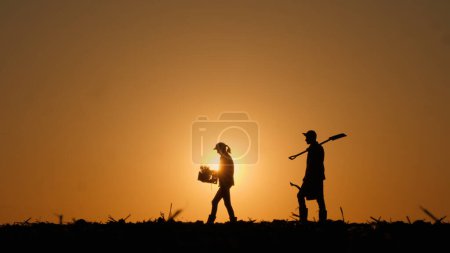 Silhouettes of two farmers walking across a field with working equipment. Side view