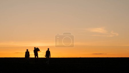 Silhouetten von drei Bauern auf einem Feld. Wandern mit Ausrüstung vor dem Hintergrund eines malerischen Sonnenuntergangs.