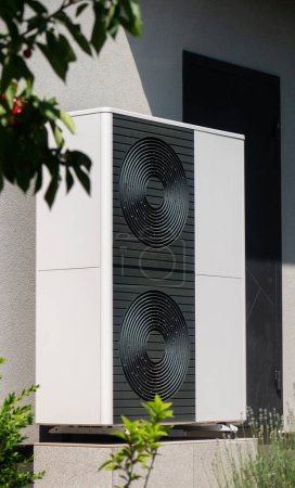 An outdoor unit of a heat pump is installed by the wall of a house. This modern and energy-efficient heating solution is neatly positioned on a concrete platform. The setup showcases the integration