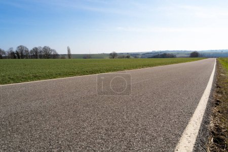 Low angle view of a paved rural road between grassfields in the landscape with blue sunny sky in spring