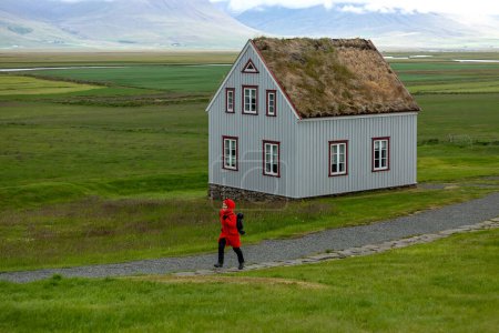 Téléchargez les photos : A girl in a red jacket runs near a peat-roofed house in Iceland. Glaumbaer - en image libre de droit