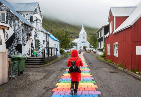 Tourist in a red coat walks along the Rainbow Road in the village of Seydisfjrur, Iceland, Scandinavia, Europe