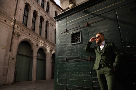 A man in glasses and a green suit stands on the street in Venice