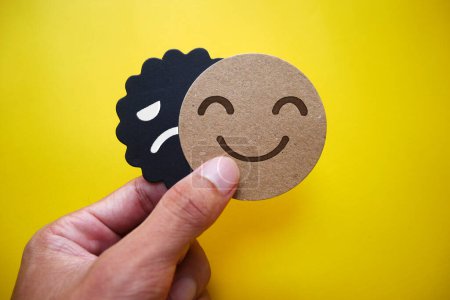 Photo for Hand holding card sad face hiding behind happy face with copy space. Concept of introversion, bipolar, depression, mental health, split personality, mood swings. - Royalty Free Image