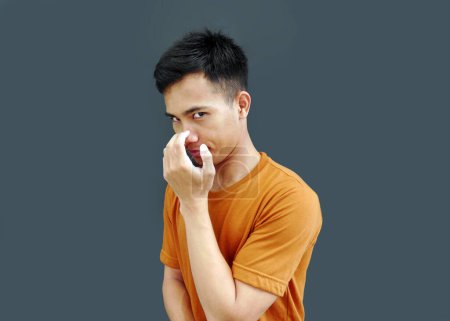 Young Asian man standing aside holding his nose and looking at the camera with an evil and cynical look.  Concept of negative human emotion, facial expressions, feelings