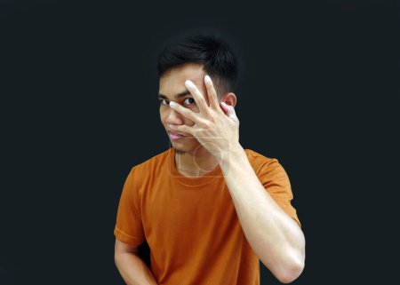 Junger asiatischer Mann bedeckt Augen mit Handflächen und guckt durch Finger, sein Gesicht sieht zynisch und scharf aus.