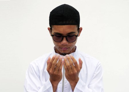 Ein junger muslimischer Mann in malaiischer Kleidung und Brille betet. Hände zum Gebet erheben. Ramadan 2024 ist voller Begeisterung.