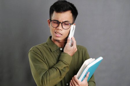 Asiático masculino estudiante en casual ropa usando gafas parece ocupado haciendo una llamada telefónica mientras lleva un libro