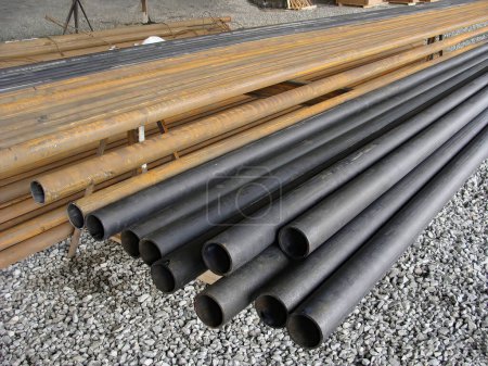 Lagerung von langen Kunststoff- und Stahlrohren auf der Baustelle, Schotterboden. Baustoffe und Strukturen.