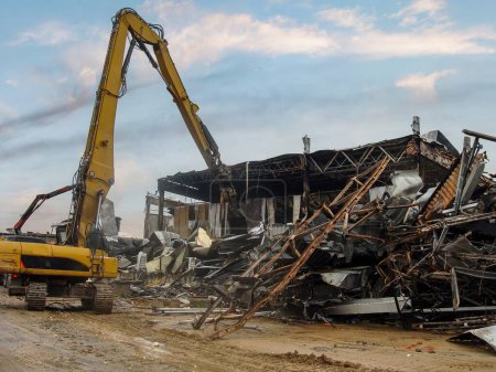 Foto de Excavadora desmontando los restos carbonizados de un almacén quemado, estructuras metálicas retorcidas visibles. - Imagen libre de derechos