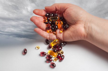 A human hand is holding a handful of various vitamins against a backdrop of a cloudy sky. The hand grips the pills firmly, showcasing the importance of health and wellness.