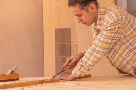 Photo for Side view of attentive male woodworker carving wood with rasp near jack plane on workbench while working in modern workshop against boards of lumber - Royalty Free Image