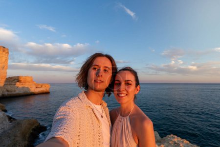 Téléchargez les photos : Golden Hour Romance : Gros plan, grand angle, prise de vue horizontale d'un couple prenant un selfie sur une falaise surplombant l'océan au coucher du soleil - en image libre de droit