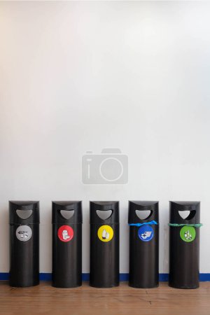 Photo for Frontal view of five black recycling bins aligned against a white wall with different colored label indicating the type of waste its meant for - general, plastic, metal, paper, and glass. - Royalty Free Image