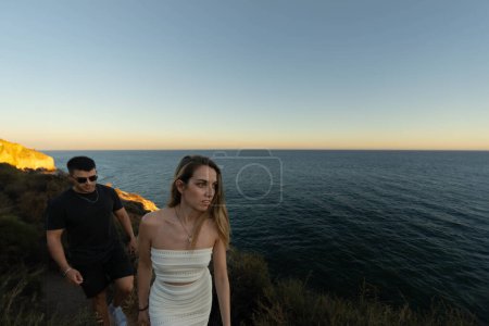 Téléchargez les photos : Plan grand angle de jeunes hommes et femmes marchant sur une falaise surplombant l'océan au coucher du soleil. - en image libre de droit