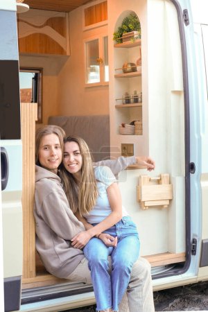Two young women relax in the door of a luxury campervan, enjoying their travel experience.