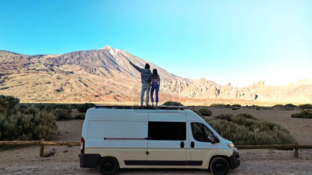 Two individuals standing on top of a van in the desert, as captured from a drone view in the Teide volcano mountain, Tenerife.